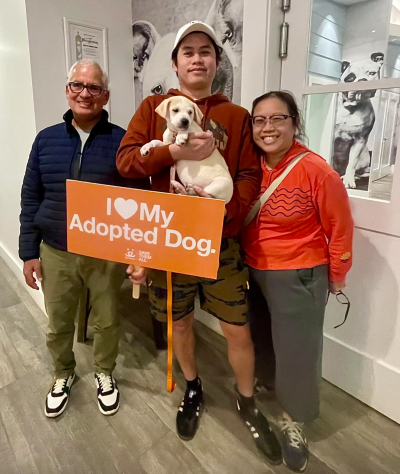 Smiling family holding a white puppy and a sign that says I heart my adopted dog